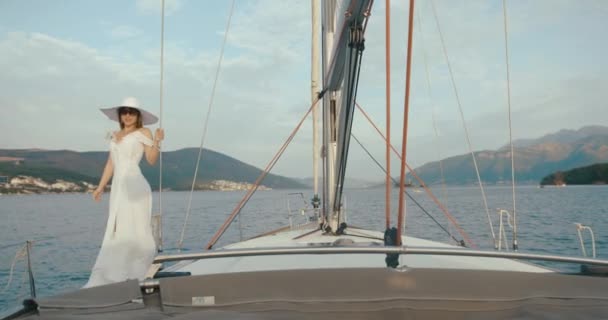 Пані на яхті. Жінка в довгій білій сукні і великий капелюх стоїть на човні, що їде через озеро у вітряну погоду — стокове відео