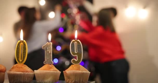 Fête du Nouvel An. Cupcakes avec des bougies en chiffres 2019 debout sur la table devant une entreprise joyeuse célébrant le Nouvel An avant un arbre de Noël — Video