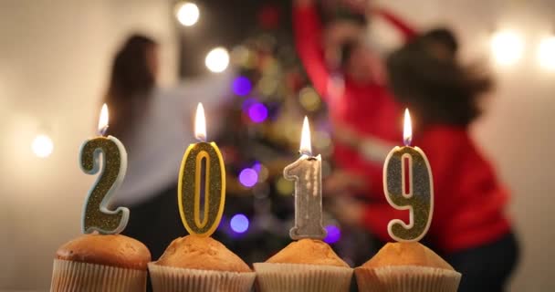 New Year celebration. Cupcakes with candles in numbers 2019 stand on the table before a cheerful company celebrating New Year before a Christmas tree — Stock Video