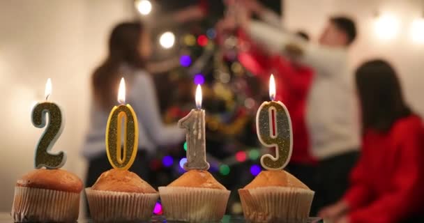 Fête du Nouvel An. Cupcakes avec des bougies en chiffres 2019 debout sur la table devant une entreprise joyeuse célébrant le Nouvel An avant un arbre de Noël — Video