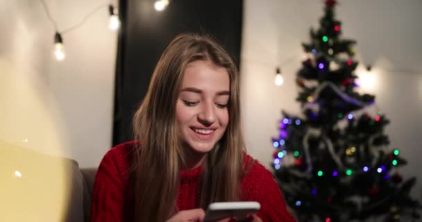 New Year celebration. Charming young woman in red sweater sits on the couch before a Christmas tree and chat with someone on her phone — Stock Video