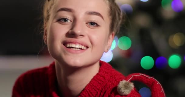 Новогодний праздник. Очаровательная женщина в красном свитере сидит на диване перед елкой, улыбается и смотрит прямо в камеру — стоковое видео