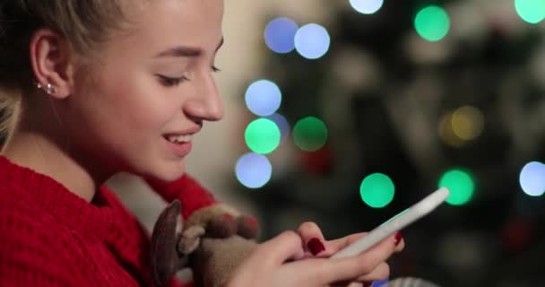 Celebração de Ano Novo. Jovem encantadora em camisola vermelha senta-se no sofá antes de uma árvore de Natal e conversar com alguém em seu telefone — Vídeo de Stock