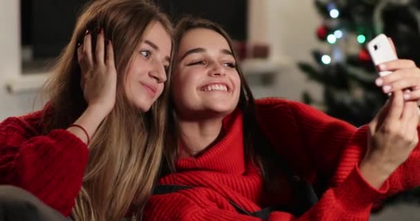 Nyårsfirande. 4 k. två tjejer ta selfie på smartphone sitter i soffan innan en julgran — Stockvideo