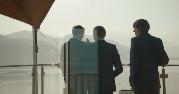 Bruiloft in Montenegro. Traditionele Europese ceremonie. Ochtend voor de receptie. 4 k. zakenlieden. Drie mannen in blauwe pakken drinken whisky staande op het balkon met prachtig uitzicht van de berg — Stockvideo