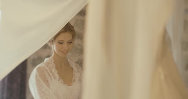 Γάμος στο Μαυροβούνιο. Παραδοσιακή ευρωπαϊκή τελετή. Το πρωί πριν από την υποδοχή. 4 k. νύφη στο λεπτό μετάξι ρόμπα θαυμάζει her γαμήλιο φόρεμα που στέκεται σε ένα δωμάτιο ξενοδοχείου — Αρχείο Βίντεο