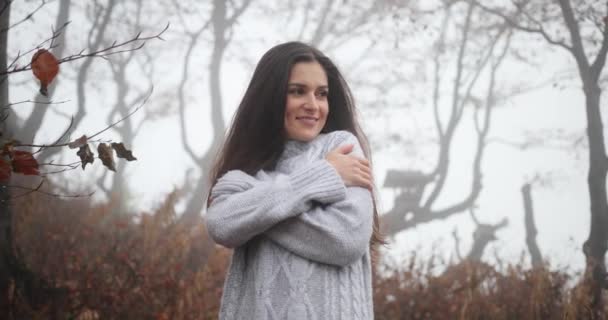 Autumn vibes, cozy atmosphere. Charming brunette woman with long dark hair embraces herself standing in the autumn forest covered with fog — Stock Video