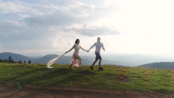 Aventura de montaña. Hombre y mujer en vestido blanco largo corren sobre la colina en los rayos del sol. 4k — Vídeos de Stock