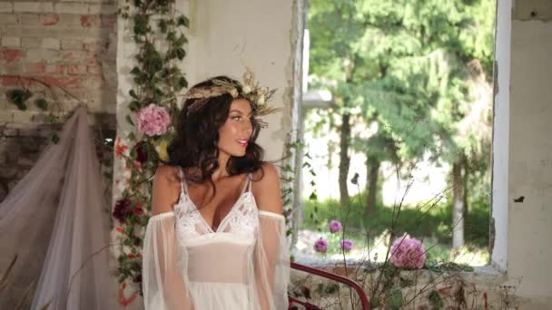 Konceptuell skytte. 4 k. kvinna med långt mörkt hår klädd som en nymf sitter på gamla säng bland blommande rosa blommor och hau och leenden tittar direkt i kameran — Stockvideo
