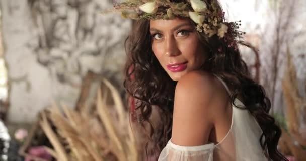 Disparo conceptual. 4k. Mujer de pelo largo y oscuro vestida como una ninfa yace en la vieja cama entre flores rosadas florecientes y hau y sonrisas mirando directamente a la cámara — Vídeos de Stock