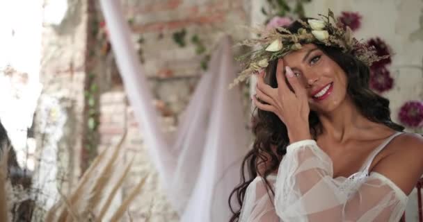 Εννοιολογική γυρίσματα. 4 κ. γυναίκα με μακριά σκούρα μαλλιά ντυμένος σαν νύμφη κάθεται στο παλιό κρεβάτι μεταξύ ανθοφορία ροζ λουλούδια και hau και χαμόγελα, κοιτάζοντας κατευθείαν στην κάμερα — Αρχείο Βίντεο