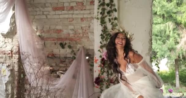 Konzeptionelles Schießen. 4k. Frau mit langen dunklen Haaren, gekleidet wie eine Nymphe, tanzt um das alte Bett zwischen blühenden rosa Blumen und Hau und lächelt direkt in die Kamera — Stockvideo