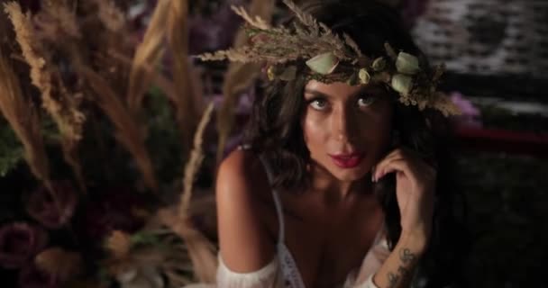 Tiroteio conceitual. 4K. Mulher com longos cabelos escuros vestida como uma ninfa senta-se na escuridão na cama velha entre flores rosa florescendo e hau e sorri olhando diretamente na câmera — Vídeo de Stock