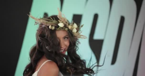 Modelo vestida como una ninfa en vestido blanco con coronas de flores baila ante una pared con letras enormes — Vídeo de stock