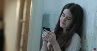 Uzun koyu saçlı gülümseyen genç kadın bir odada yerde oturan akıllı telefonda bir şey okur