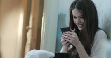 Uzun koyu saçlı gülümseyen genç kadın bir odada yerde oturan akıllı telefonda bir şey okur