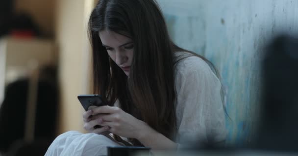 Jovem sorridente com cabelos longos e escuros lê algo em seu smartphone sentado no chão em uma sala — Vídeo de Stock