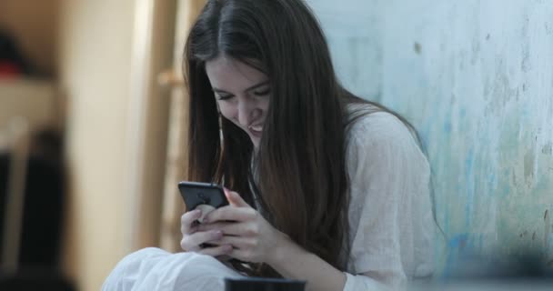 Lächelnde junge Frau mit langen dunklen Haaren liest etwas in ihrem Smartphone, das auf dem Boden in einem Raum sitzt — Stockvideo