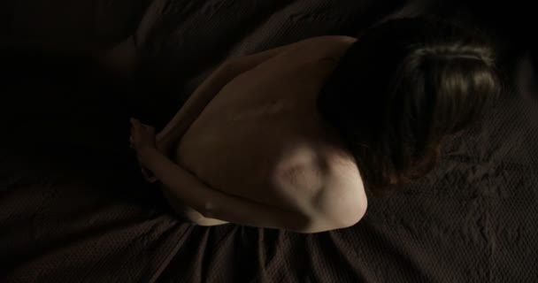 Η ιδέα του θύματος. Νέα κοκαλιάρικο μελαχρινή γυναίκα με σκούρα μαλλιά κρατά τα χέρια της πίσω από την γδαρμένη πίσω κάθεται στο κρεβάτι — Αρχείο Βίντεο