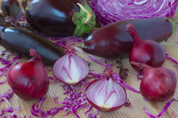 Two heads of purple onion, the two halves of onion, eggplant and purple basil and sliced purple cabbage on the tabl