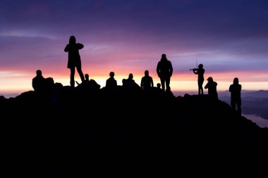 Çok renkli gün batımı. Kemanlı bir kadının silueti. Denize bakan bir dağın tepesinde insanların siluetleri