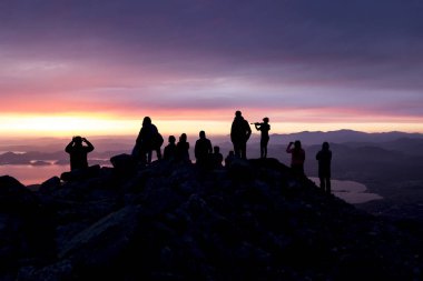 Çok renkli gün batımı. Kemanlı bir kadının silueti. Denize bakan bir dağın tepesinde insanların siluetleri