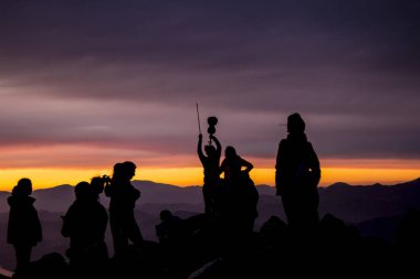 Çok renkli gün batımı. Kemanlı bir kadının silueti. Denize bakan bir dağın tepesinde insanların siluetleri