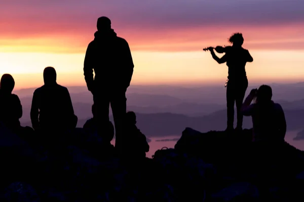 Tramonto multicolore. Silhouette di una donna con un violino. Silhouette di persone sulla cima di una montagna con vista sul mare — Foto Stock