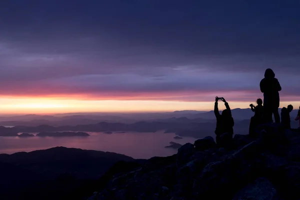 Puesta de sol multicolor. Siluetas de personas en la cima de una montaña con vistas al mar —  Fotos de Stock