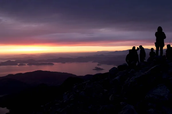 Puesta de sol multicolor. Siluetas de personas en la cima de una montaña con vistas al mar —  Fotos de Stock