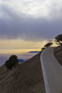Dağlardaki yol, dağların yamaçlarındaki ağaçlar, sarı gün batımı, bulutlardaki gökyüzü ve deniz