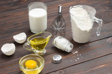 Unlu ölçü kabı, bir bardak süt, kırık yumurta ve tuz, masada metal çırpı. Koyu ahşap arka plan. Üst görünüm