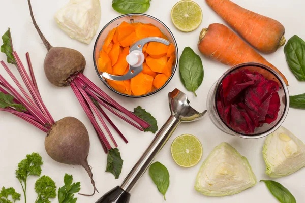 Chopped beets in jar, chopped carrots in blender jar. Beets with tops and unpeeled carrots. Metal shredder. Cabbage and lemon halves, basil and parsley. Flat lay. White background. Copy space