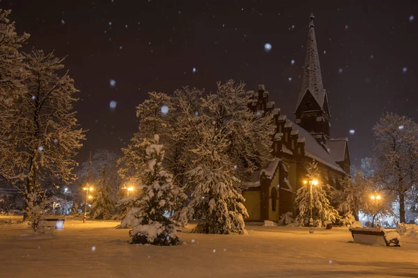 Buzlu bir akşam donmuş ve kapalı kar ağaçları ile çevrili bir kış parkı eski bir kilise ile güzel bir manzara. Küçük parkta kar yağışı. Arka planda eski kilise ve kar altında Ağaçlar.