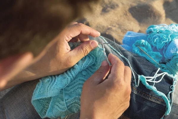 man\'s hands with knitting, needles and knitted thing in process of knitting, next to the knitter is blue yarn