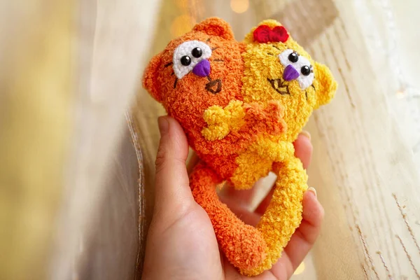 toy knitted two red and yellow love cats made of wool tails connect in the heart against background of n female palm against background of yellow airy fabric, , handmade, copy space