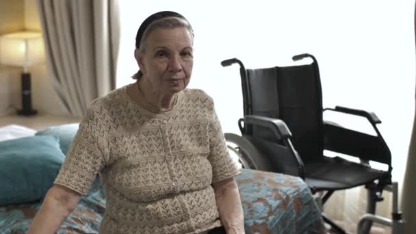 Staruszka siedzi na łóżku obok wózka inwalidzkiego — Wideo stockowe
