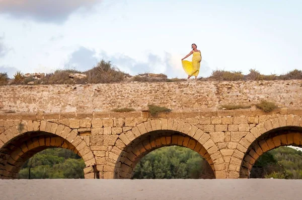 Young and attractive Caucasian pregnant woman in a long yellow dress dancing on the top of ancient ruins - Roman aqueduct. Expecting a baby, giving birth to a child, natural birth concept, awaiting a child.