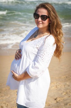 Onun geç 20s onun göbek tutarak mutlu güzel genç hamile Kafkas Rus kadın, o onun arka planda deniz dalgaları ile deniz kenarında sıcak bir yaz gününde güneş gözlüğü ile bir hasır şapka giyiyor. Sahilde hamile genç kadın, tatil