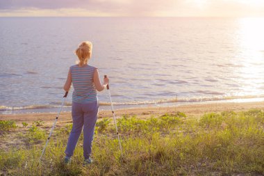 Güzel genç kız sarışın sopa hiking ile kolları olmadan çizgili gömlekli günbatımında görünüyor. Arkadan görünüm