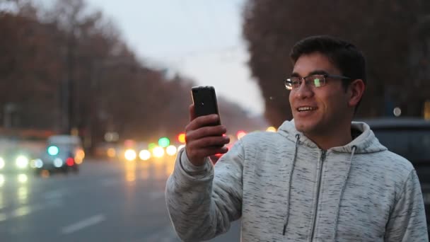 Un ragazzo con gli occhiali e una camicia bianca parla su skype con il telefono nero e ride, Su uno sfondo bokeh dei fari auto in movimento . — Video Stock