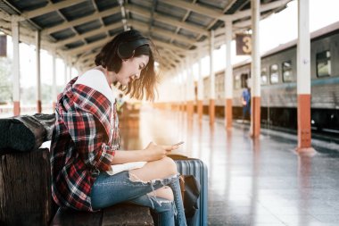Gezgin kız oturma ve tren istasyonunda bekleyen bir Bagaj ile Tablet ve kulaklık kullanarak. Açık sergüzeşt seyahat etmek tren konsepti vintage filtre ile tarafından