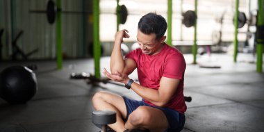 Kırmızı tişörtlü bir adam dirseğini tutarken yüzünü buruşturuyor. Spor salonunda antrenman yaparken rahatsız olduğunu gösteriyor..