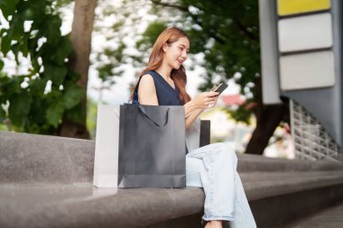 Rahat bir kadın, dışarıda bir bankta akıllı telefonunu kontrol ederken alışveriş deneyiminin tadını çıkarıyor..