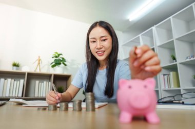 Bir kadın, pembe bir kumbarada para biriktirirken, finansal planlama ve yetkilendirmeyi temsil eden bir not defterine yazıyor..