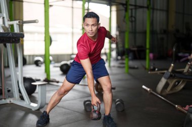 Kırmızı tişörtlü bir adam kettlebell egzersizi yapar. Gücünü gösterir ve spor salonuna odaklanır..