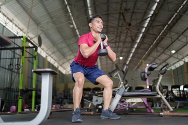 Kişi, elinde bir dambıl tutarken çömelir, spor salonunda kuvvet eğitim tekniklerini vurgular..