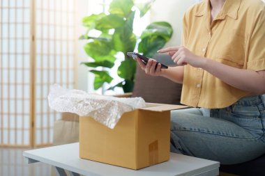 Bir kadın akıllı telefonunu kontrol ederken heyecanlı bir şekilde bir teslimatı açıyor ve online alışverişin keyfini çıkarıyor..