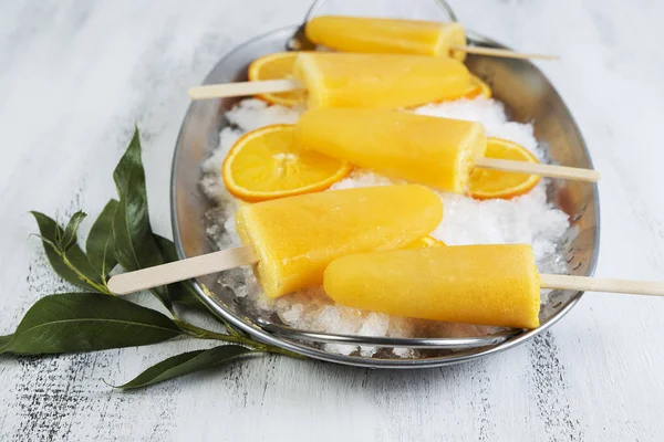 Homemade orange frozen juice on a stick on ice on a light wooden background.