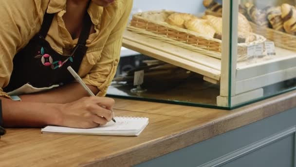有吸引力的成熟妇女工作她的面包店做笔记在笔记本上愉快地微笑对照相机女实业家零售购买消费主义食品糕点烘焙休闲产业 — 图库视频影像